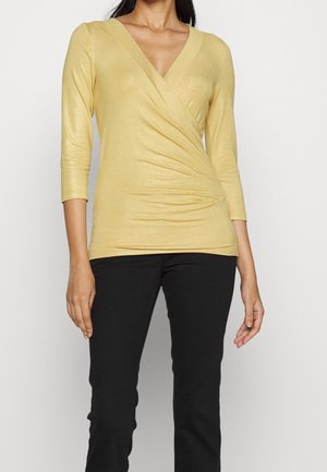 Kvinde iført en gul slå-om-top med trekvartærmer og sorte bukser, stående mod en ensfarvet hvid baggrund.