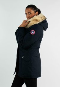 Parka marine avec capuche doublée en fourrure, dotée d'un patch logo rouge et bleu, d'un système de fermeture à boutons et d'un design légèrement cintré.
