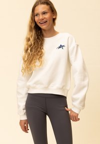 Witte sweatshirt met geribbelde manchetten en zoom, met een meerkleurig sterrenlogo op de borst. Draagt grijze leggings. Soepele textuur.