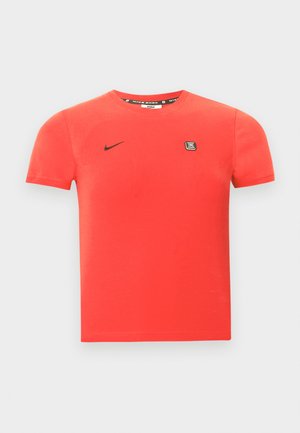 Raudonas medvilninis marškinėlis trumpomis rankovėmis, su juodu „Nike“ logotipu ir mažu firminiu lopu ant krūtinės. Paprastas apvalus kaklas.