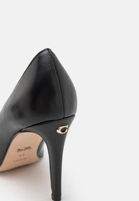 Chaussure à talon haut en cuir noir avec bout pointu et détail décoratif doré sur le talon, sur fond blanc.