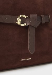 Pruun suede käekott, millel on nahkrihm kuldse ümmarguse lukuga ja brändinimi "Coccinelle" kuldse tähega.