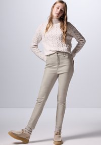 Beige high-waisted broek met voorzakken, gecombineerd met een crèmekleurige gebreide trui en lichtbeige schoeisel.