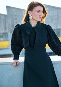 Femme aux cheveux roux portant de grandes boucles d'oreilles argentées et une robe noire à manches bouffantes, appuyée sur un rebord en béton à l'extérieur.