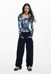 Haut à manches longues et ajusté de couleur bleue avec des motifs abstraits, associé à un pantalon cargo ample bleu marine. Le mannequin porte un collier avec un pendentif en forme de cœur argenté.