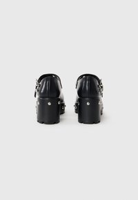 Black heeled shoes with a smooth leather exterior, a chunky platform sole, silver stud accents, and an adjustable strap at the heel.