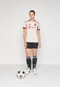 Hvid fodboldtrøje med korte ærmer og røde detaljer og logo, parret med sorte shorts og knælange sokker, sammen med en sort og hvid fodbold.