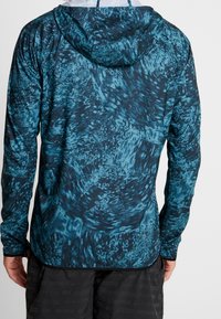 Haut à capuche à manches longues en bleu foncé avec des motifs tourbillonnants en bleu plus clair. Tissu lisse et léger avec une coupe décontractée et des trous pour les pouces aux poignets.