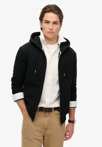 Schwarze Zip-Hoodie mit zwei Vorderaschen, Kordelzugkapuze und gerippten Bündchen. Über einem weißen, knöpfbaren Hemd mit navyfarbenem Akzent getragen.
