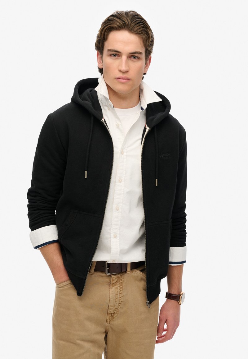 Schwarze Zip-Hoodie mit zwei Vorderaschen, Kordelzugkapuze und gerippten Bündchen. Über einem weißen, knöpfbaren Hemd mit navyfarbenem Akzent getragen.