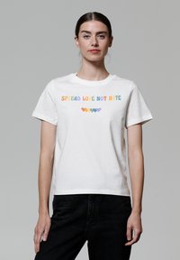 T-shirt en coton blanc affichant la phrase "RÉPANDRE L'AMOUR PAS LA HAINE" en lettres multicolores avec des icônes de cœur en dessous. Manches courtes, coupe classique.