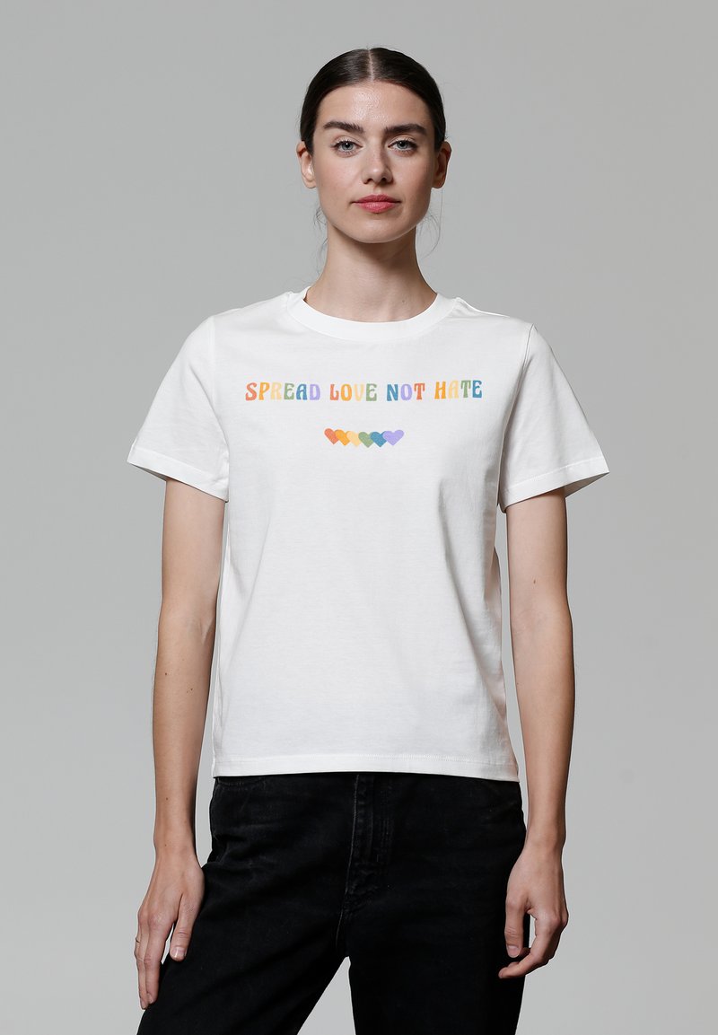 T-shirt en coton blanc affichant la phrase "RÉPANDRE L'AMOUR PAS LA HAINE" en lettres multicolores avec des icônes de cœur en dessous. Manches courtes, coupe classique.