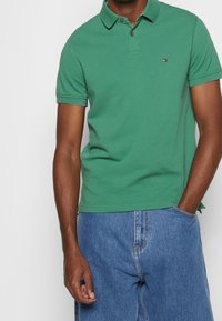 Green polo shirt made from cotton with a collar and two buttons, featuring a small logo at the chest. Paired with blue denim shorts.