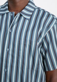 Camisa de manga corta con botones en azul, verde y marrón con rayas verticales. Presenta un bolsillo en el pecho y un cuello puntiagudo. Textura de tela suave.