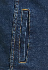 Denimová kapsa v tmavě modré barvě, s vertikálním švem v kontrastní oranžové nitce. Hladký povrch a čisté okraje.