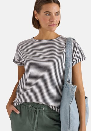 Femme portant un t-shirt rayé noir et blanc, un pantalon vert à cordon et un sac cabas bleu clair à l'épaule.