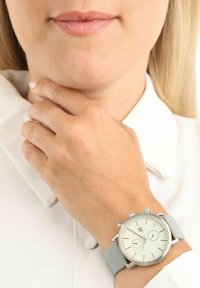 Silberne Armbanduhr mit einem weißen Zifferblatt, grauem Lederarmband, drei kleinen Ziffernblättern und silberfarbener Hardware, getragen an einer hellhäutigen Hand vor einem weißen Hintergrund.