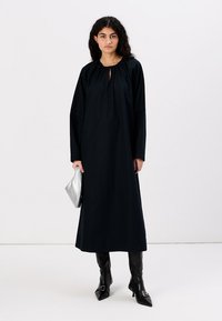 Lang svart kjole med en nøkkelhull-halsutskjæring og voluminøse lange ermer. Modellen holder en sølvfarget clutch og har på seg svarte høyhælte støvler til knærne.