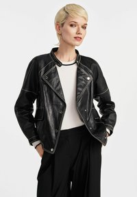 Czarna skórzana kurtka z białym przeszyciem, z ząbkowanym kołnierzem i zatrzaskami. Nosi ją osoba w białej bluzce, w połączeniu z czarnymi spodniami.