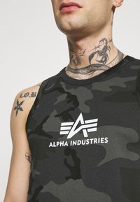 Maskovací tielko v tmavozelenej a čiernej farbe, s bielym logom "Alpha Industries", okrúhlym výstrihom a bezrukávovým dizajnom.