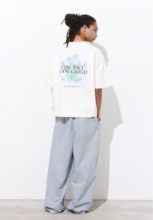 Personne avec des dreadlocks de profil portant un T-shirt blanc oversize avec le texte « Vincent Van Gogh » et un pantalon large à carreaux bleu clair.