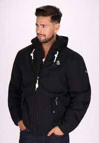 Giacca nera con zip e cappuccio, con lacci bianchi e logo sul petto. Realizzata in tessuto liscio, con tasche laterali con zip e polsini elasticizzati.