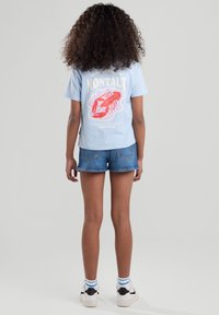 T-shirt di cotone azzurro chiaro con grafica di aragosta e testo. Abbinata a pantaloni corti in denim. Modella in piedi, vista da dietro. Sneakers bianche e nere.