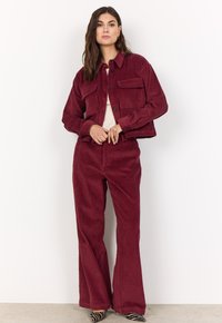 Burgundy corduroy jacket with two chest pockets and black buttons, paired with matching corduroy flared pants and striped heels.
