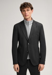 Herren maßgeschneiderter dunkelgrauer Blazer mit zwei vorderen Taschen, silberner Knopfverschluss, übereinander getragen mit einer hellgrauen Zip-Jacke.