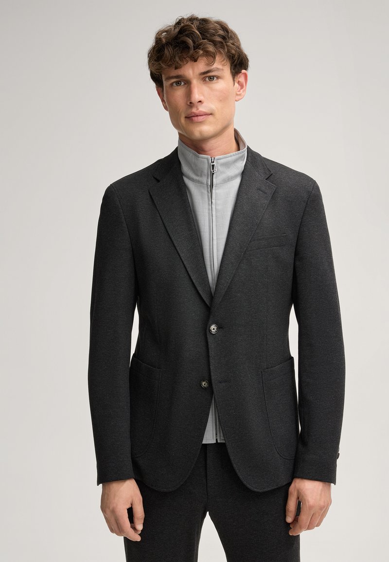 Herren maßgeschneiderter dunkelgrauer Blazer mit zwei vorderen Taschen, silberner Knopfverschluss, übereinander getragen mit einer hellgrauen Zip-Jacke.