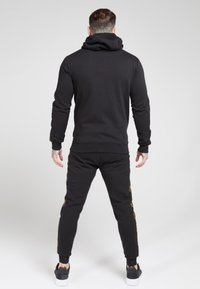 Completo felpa con cappuccio e pantaloni in nero, caratterizzato da un cappuccio e dettagli laterali dorati sui pantaloni, realizzato in materiale morbido ed elastico.