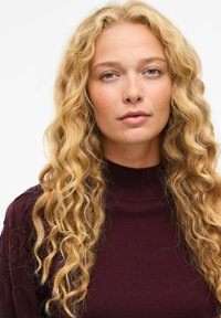 Mörkröd polotröja med ribbad textur, som har långt, vågigt blont hår som faller ner, neutral bakgrund som framhäver plagget.
