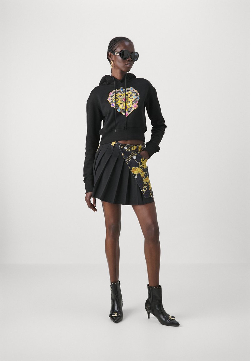 Sweat à capuche noir court avec un motif floral coloré, associé à une jupe plissée noire avec des accents dorés. Des bottines noires complètent la tenue.