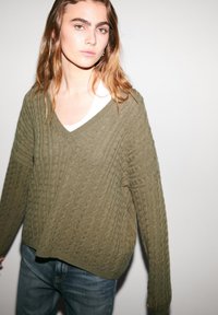 Olive green cable-knit sweater with a v-neckline and long sleeves, paired with a white undershirt, worn over blue jeans.