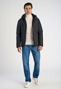 Mörk vadderad jacka med huva, bärs över en beige polotröja, ihop med blå jeans och vita sneakers. En enkel bakgrund.