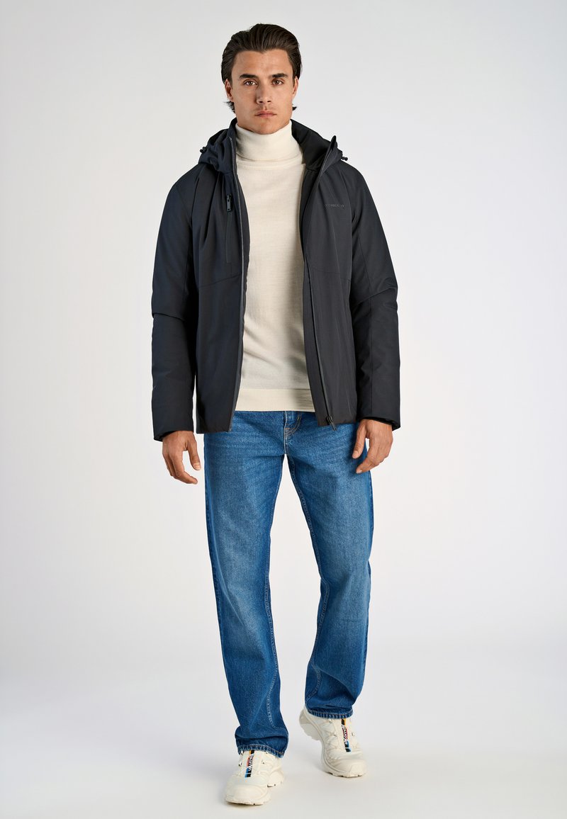 Mörk vadderad jacka med huva, bärs över en beige polotröja, ihop med blå jeans och vita sneakers. En enkel bakgrund.