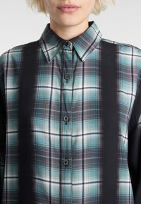 Chemise à carreaux à devant patte de boutonnage avec un motif en plaid noir et bleu canard, équipée d'un col pointu et de deux boutons au niveau du col.