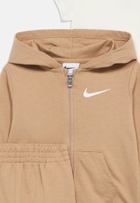 Hnedá mikina na zips s kapucňou a elastickým pásom, zdobená bielym logom Nike. Mäkká tkanina, kombinovaná s krémovým tričkom pod ňou.