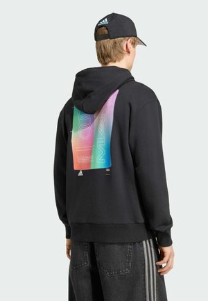 Henkilö, jolla on päällään musta huppari, jossa on värikäs pystysuuntainen kuvio selässä, ja musta Adidas-lippis, selin kameraan.