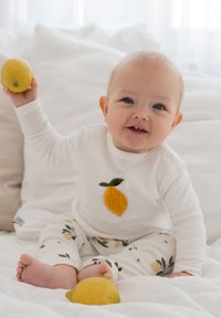 Weißes Langarmhemd mit einem gelben Zitronenmotiv, kombiniert mit weißen Hosen, die mit grünen Blättern und Zitronenmuster verziert sind. Baby hält eine Zitrone.