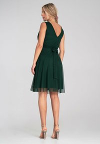 Grünes ärmelloses Kleid mit V-Rücken, tailliert, mit einem geschichteten Tüllrock und glattem Stoff. Es werden klare High-Heel-Schuhe getragen.