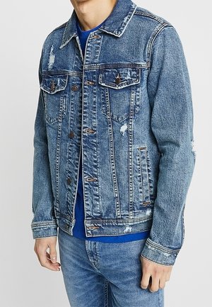 Veste en denim bleu de coupe classique, avec des accents déchirés, deux poches poitrine et des détails en métal. Portée sur une chemise bleue.