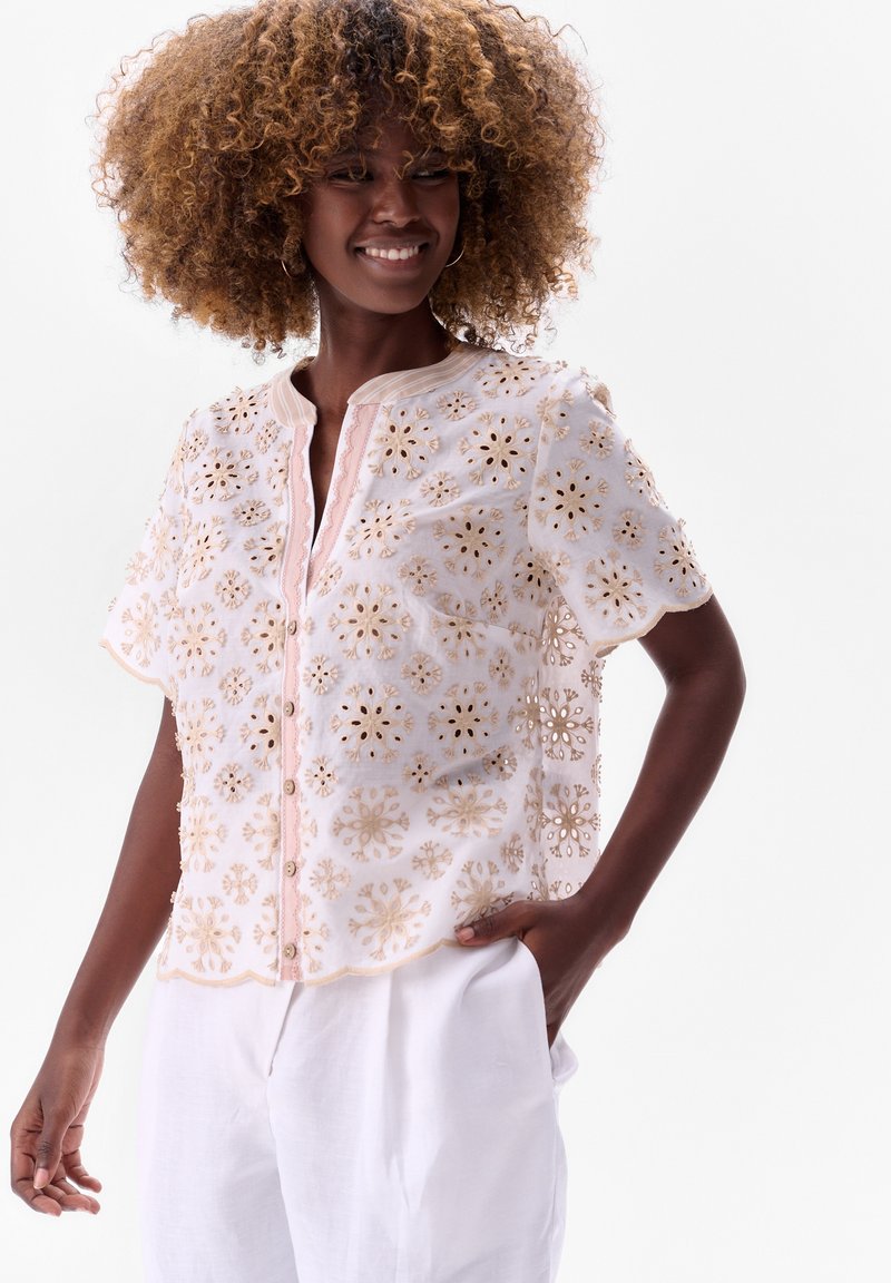 Witte geborduurde blouse met bloemenpatronen, gouden accenten, korte mouwen, een ronde halslijn en een knopenrij aan de voorkant. Gecombineerd met witte broeken.