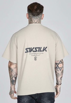 Homme avec tatouages sur le cou et le bras, portant un t-shirt beige SikSilk avec un logo noir et du texte, vu de dos sur un fond gris uni.