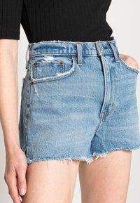 Pantalones cortos de denim azul claro con dobladillos deshilachados, de cintura alta, cinco bolsillos, remaches de cobre y un ligero desvanecimiento. El material parece robusto pero flexible.