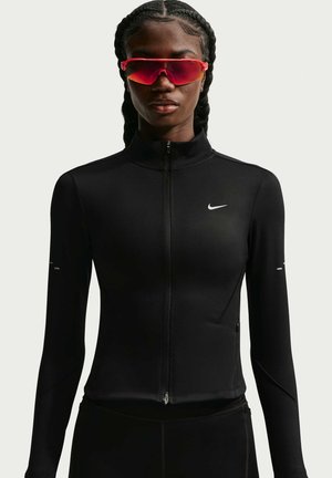 Atleet draagt een zwart Nike jack met ritssluiting en rode sportzonnebril, met gevlochten haar tegen een effen achtergrond.