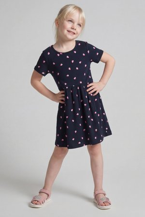 Mladé dievča so slovenskými vlasmi, oblečené v námorníckych šatách s ružovým motívom jahôd a ružovými sandálmi, stojace s rukami na bokoch, usmievajúce sa.