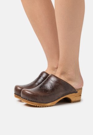 Sabots en cuir brun avec une finition texturée, dos ouvert, semelle en bois et coutures surélevées. Le design présente des lignes courbes et une pointe arrondie.