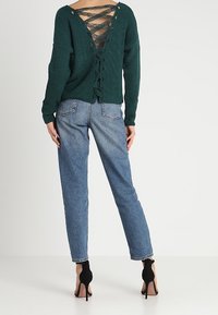 Pull en maille vert foncé avec un dos décolleté orné de lacets croisés. Associé à un jean bleu clair et des chaussures à talons noires.