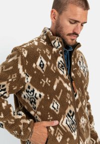Braune Fleecejacke mit einem geometrischen Muster in Creme und Schwarz. Sie hat einen hohen Kragen und einen Reißverschluss, sowie Seitentaschen für Stauraum.